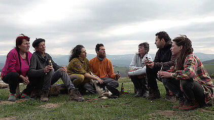 Sept bergers sont assis en arc de cercle dans un champs sur les hauteurs de l'Ariège, ils discutent et trinquent après la transhumance